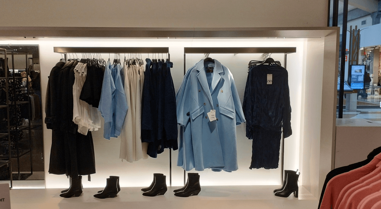 A retail clothing display showcasing neatly arranged garments. The rack features a range of outfits, including black and white tops, light blue shirts, dark blue dresses, and a prominent light blue double-breasted coat with a price tag. Below the clothing, black ankle boots are aligned on a well-lit shelf. The display is modern, with a sleek and minimalist design, situated near a shopping mall corridor.
